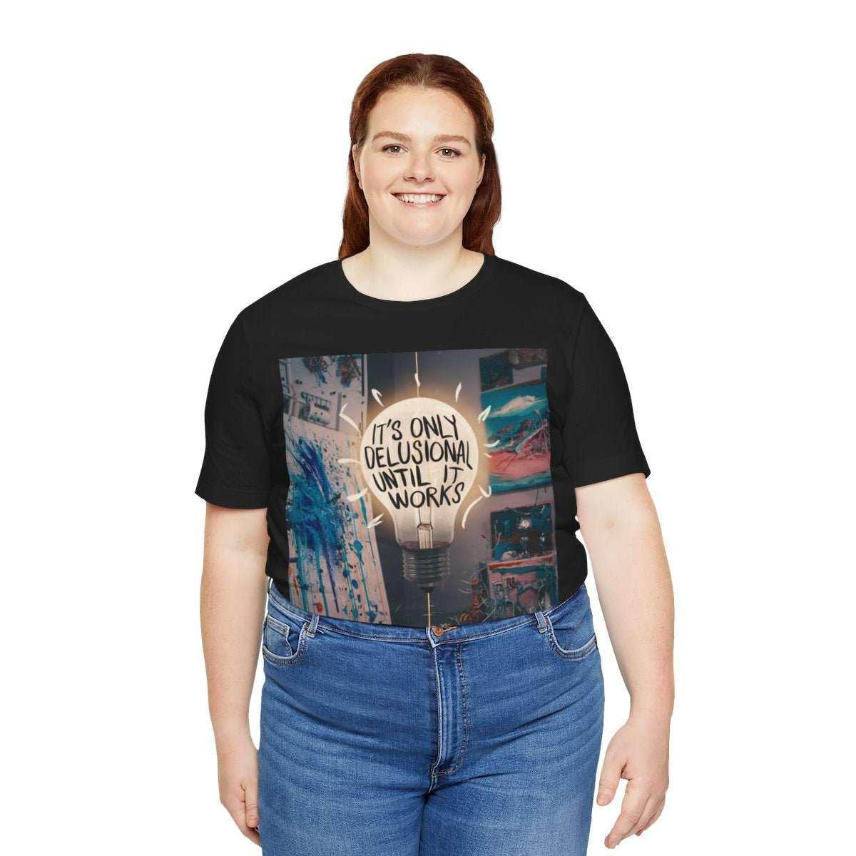 Smiling woman wearing black graphic tee with 'It's only delusional until it works' lightbulb design.