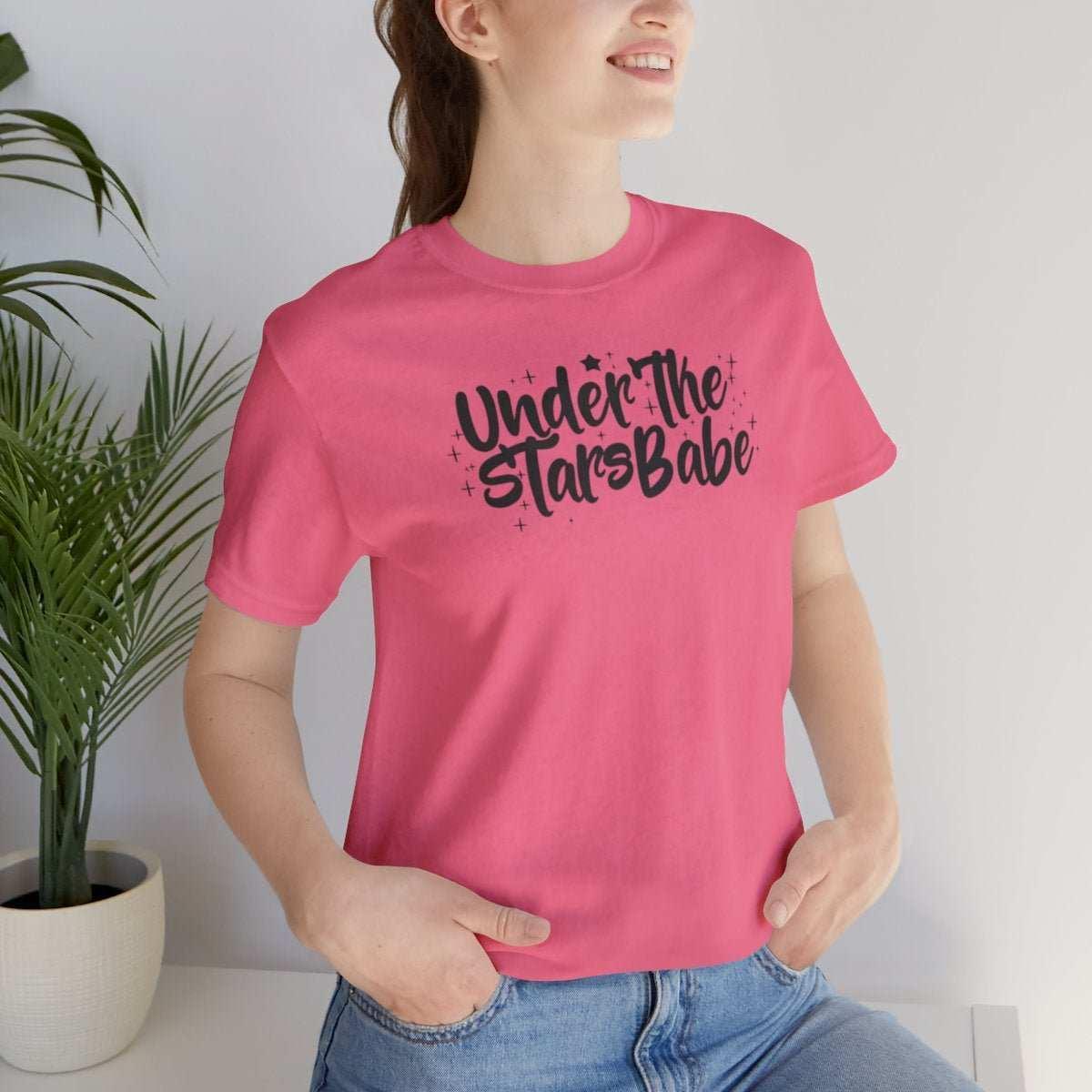 Woman in pink Under the Stars Babe t-shirt and jeans, standing indoors near plants